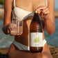 Aloe Vera Saft eingeschenkt im Glas, neben Wild Baboon Glasflasche wird von Mädchen im weißen Bikini gehalten