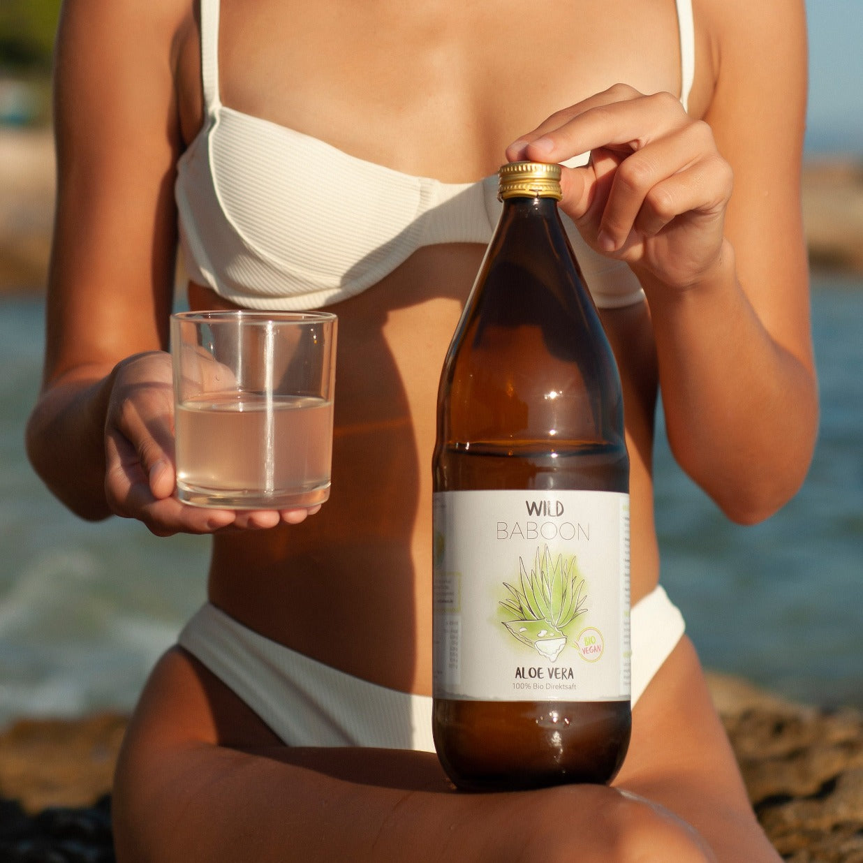 aloe vera gel trinkt Frau im Bikini und hält Wild Baboon Flasche in der Hand